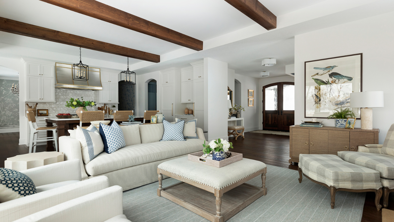 an open concept kitchen and living room with a neutral color scheme and blue accent decor, exposed beams on the ceiling, a couch, an ottoman, and a plaid accent chair in one corner