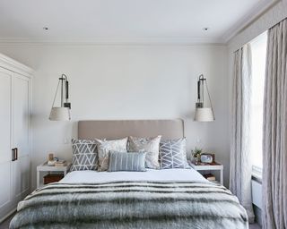 Double bed with grey and silver cushions and bed throws, built in cupboards to the left and sash window with long curtain to the right.