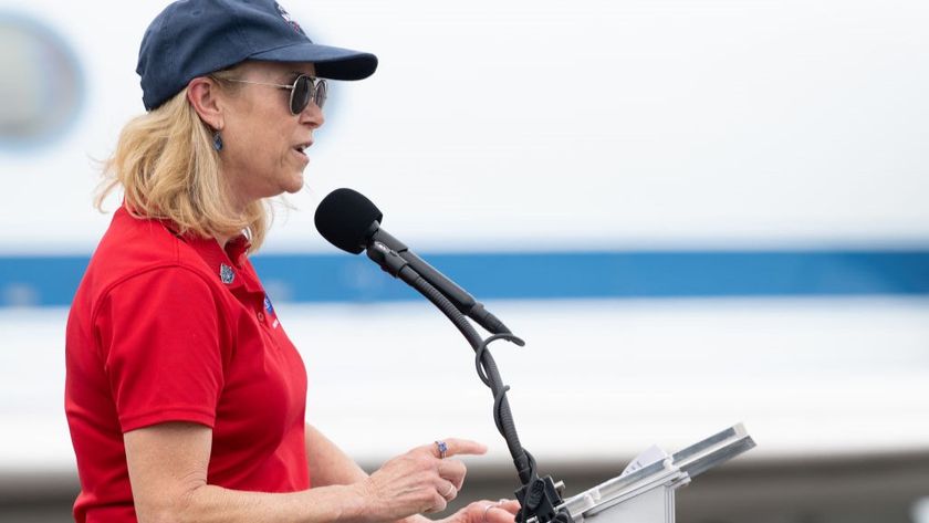 A woman with light hair is wearing a red shirt, blue hat and sunglasses, facing right, speaking into a microphone.