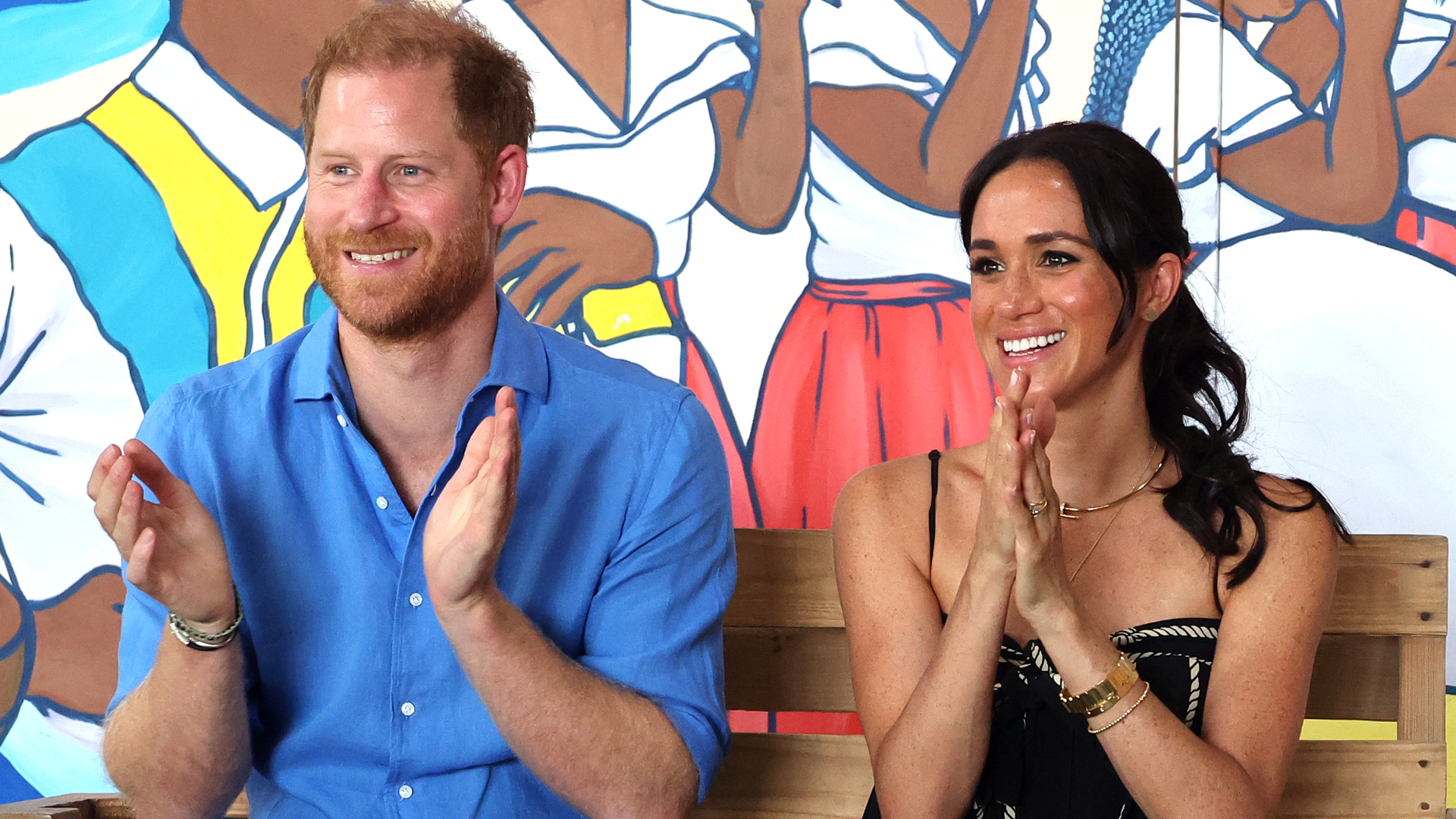 Prince Harry wears a blue shirt and brown pants and claps next to wife Meghan Markle who wears a black and white striped dress