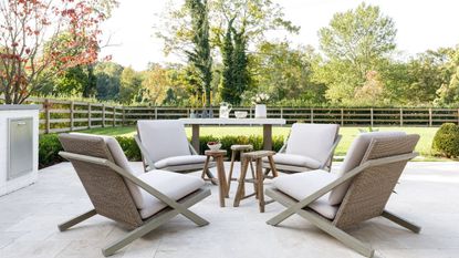 A white stone garden patio with modern white and rattan furniture. Behind is a large green lawn with wooden fencing, and tall trees are seen in the background.