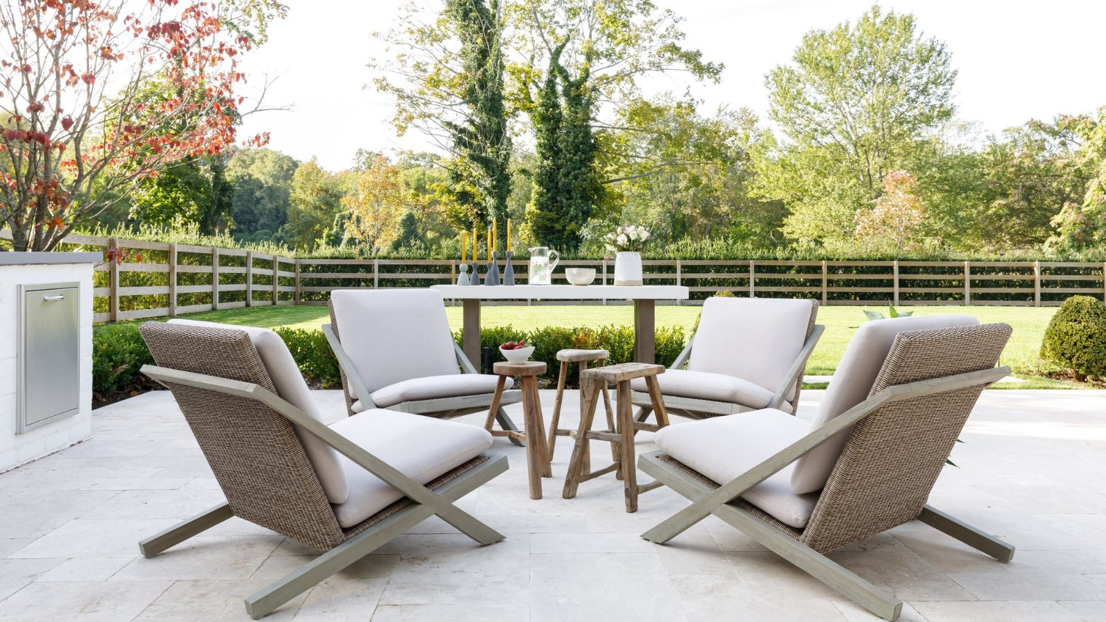 A white stone garden patio with modern white and rattan furniture. Behind is a large green lawn with wooden fencing, and tall trees are seen in the background.