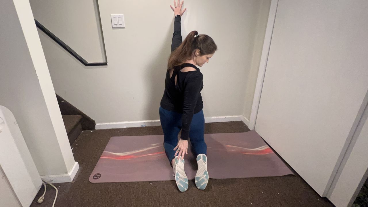 Trainer Jennifer RIzzuto performs a back mobility stretch on the floor. She is kneeling close to a wall on a yoga mat, left hand on the wall and right hand reaching back towards her left heel.