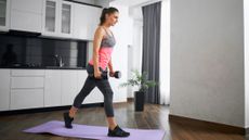 Woman stands in a kitchen on an exercise mat holding dumbbells. She stands with one leg behind her. She is sportswear, a tank top, three-quarter grey leggings, and black trainers. Her brown hair is in a pony tail.