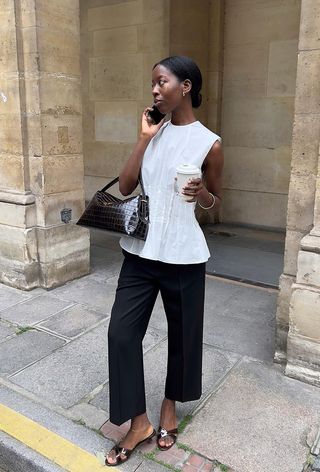 a photo showing what shoes to wear with wide-leg pants with a woman on the sidewalk wearing a white blouse with wide-leg cropped black trousers, brown open-toed mule sandals, and a brown handbag