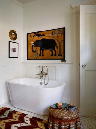 Image of a white bathroom with white tile floors and a white stand-alone bathtub. The door is ajar and there is a tartan-print, small ottoman in front of it. under the ottoman and in front of the tub is a red, orange, light pink, and sage green chevron rug.