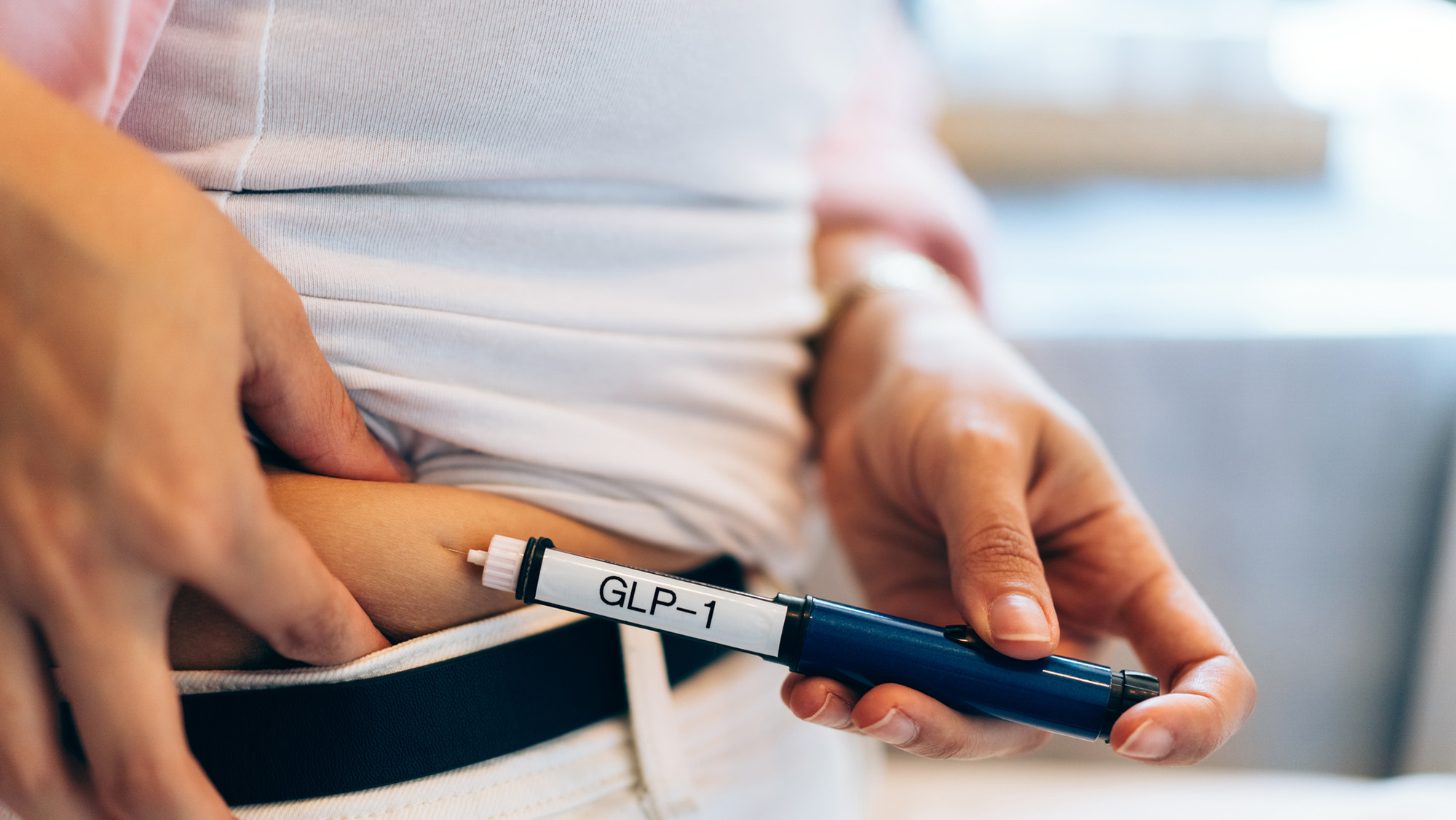 A close up of a woman injecting her stomach with a white cylinder with the label "GLP-1"
