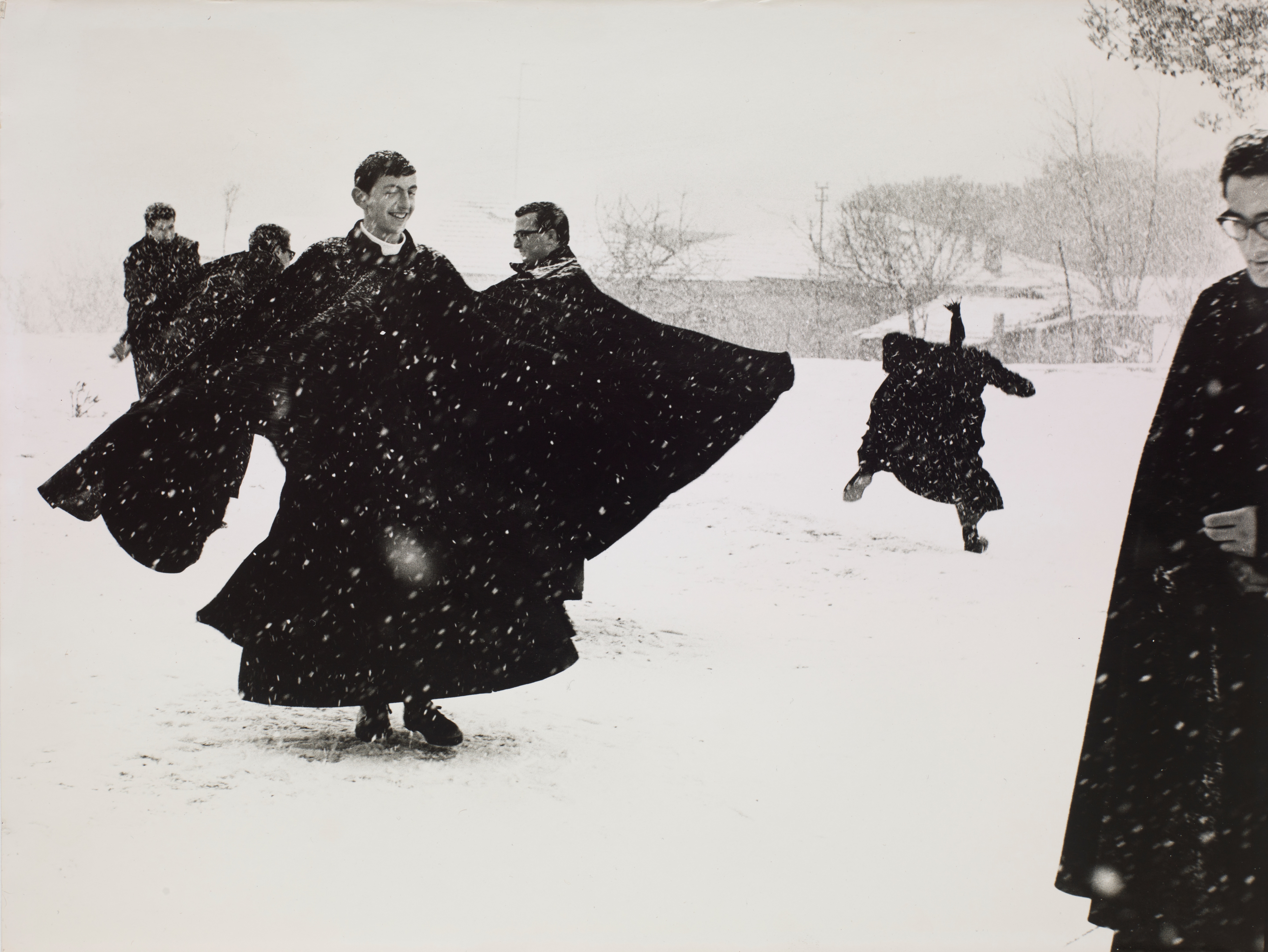 A group of young priests in flowing black cassocks caught mid-motion in falling snow, one leaping joyfully with arms outstretched.