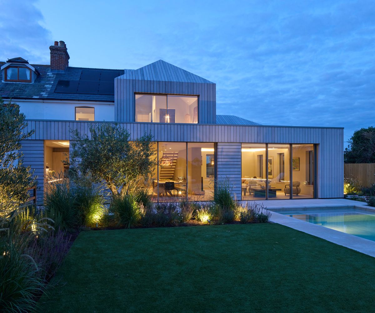 exterior shot of timber clad extension at night with double storey addition in centre of roof, gardens and swimming pool outside
