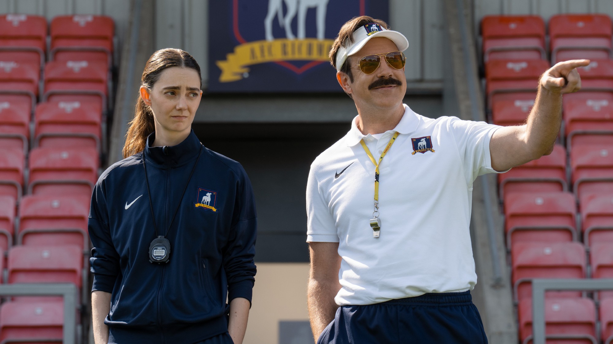 From left to right Tanya Reynolds standing with her hands in her pockets and Jason Sudeikis pointing to the right with his right hand on Ted Lasso.