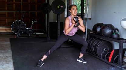 Woman performs side lunge in garage gym. She holds a kettlebell upside down by her chest. She wears black three-quarter length sports leggings, black trainers and a pink tank top.