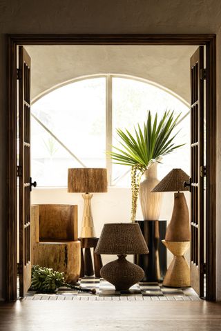 A series of wooden and rattan lamps sits in a naturally lit room past an open wooden door. Next to them are a few plants adding a touch of green to the space,