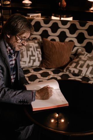 Image of Marc Newson writing in an open book in a dimly lit room, round black gloss table, brown and white pattern sofa with brown cushion, blurred surroundings in the backdrop