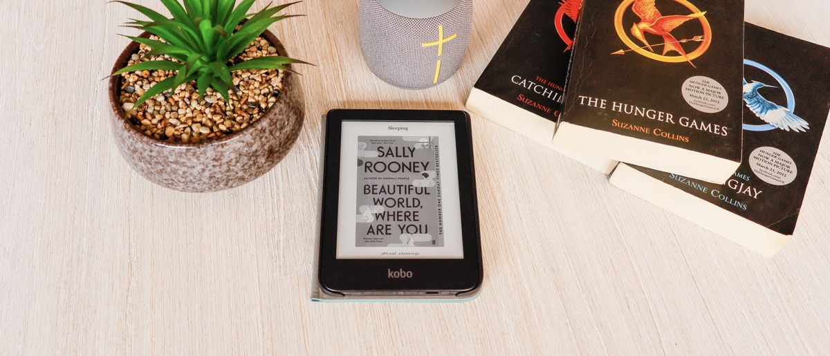 The Kobo Clara 2E laying flat on a table surrounded by a fake plant, speaker and books while showing the front cover of a book.