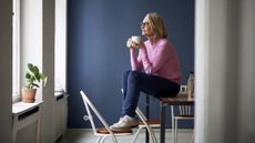 A woman sits on a table with her feet on a chair as she holds a cup of coffee and looks out the window.