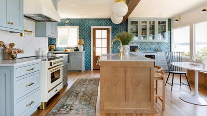 large blue kitchen with sky blue cabinetry, white countertops, brass fixtures and fittings, rug, tiled far wall, turquoise, globe pendant lights, wood island,