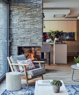 Open-plan living area with slate stone feature wall and fireplace, light wooden flooring with blue patterned rug, wooden lounge chair with upholstered gray cushions, rounded side table with geometric pattern, looking out onto kitchen with kitchen island and cupboards