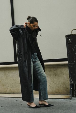 A black turtleneck outfit is shown in a photo of a French woman on the sidewalk wearing a long black leather trench coat over a black turtleneck with straight-leg jeans and black heeled mules