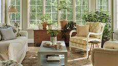 a sunny english style sunroom in connecticut with woven rug, floral armchair, lots of plants, antique wooden furniture and a large white couch
