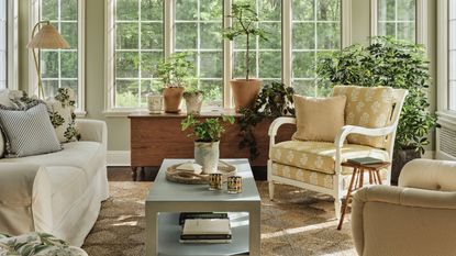a sunny english style sunroom in connecticut with woven rug, floral armchair, lots of plants, antique wooden furniture and a large white couch