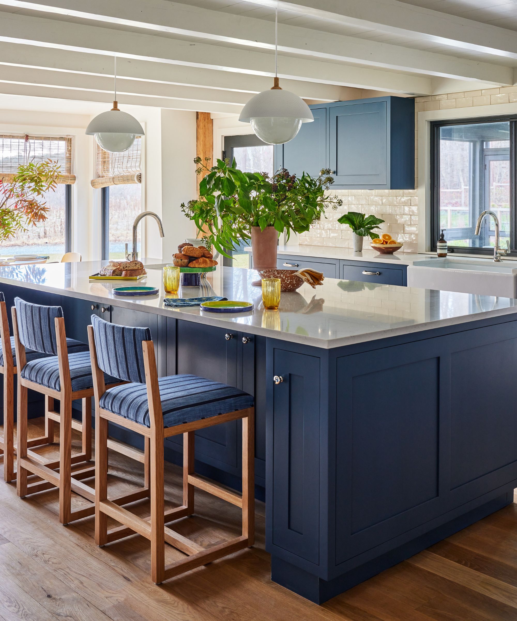 Kitchen by Sarah Lederman featuring Stiffkey Blue cabinetry, a large island with white countertops, oak flooring, and expansive windows overlooking the landscape.