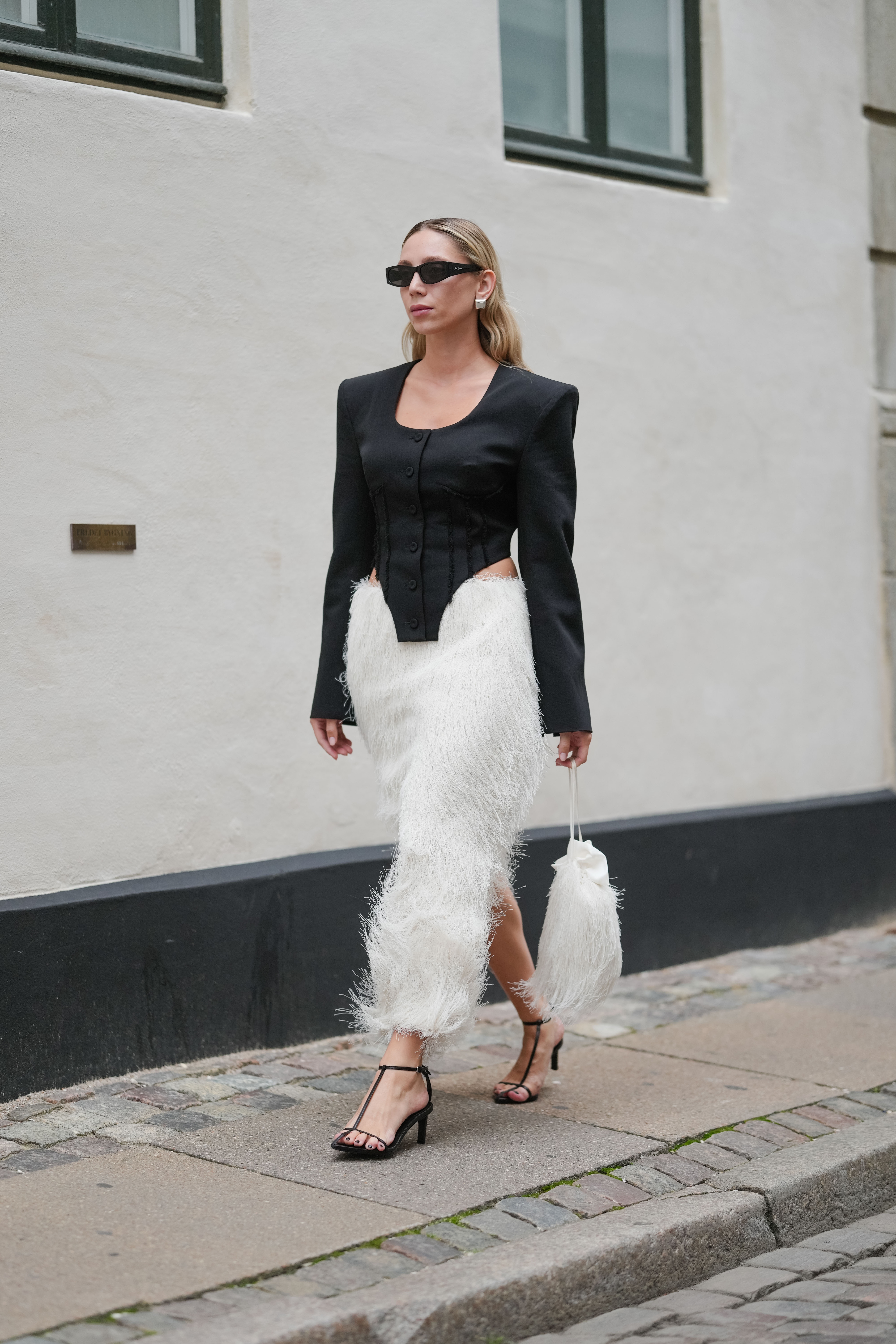 COPENHAGEN, DENMARK - AUGUST 07: A guest wears straight shoulder-length blonde hair with a center part, narrow rectangular black sunglasses, and small silver hoop earrings, a black tailored jacket in matte suiting with padded shoulders, a scoop neckline, long sleeves, corset-style seaming, multiple front buttons, a curved hem, and side cutouts at the waist, a high-waisted white midi skirt densely fringed with wispy eyelash threads, a small white satin drawstring pouch with a long cord strap and feathery trim carried by hand, and black open-toe T-strap sandals with slim mid heels and a dark pedicure, walking, outside Nicklas Skovgaard, during Copenhagen Fashion Week, on August 07, 2025 in Copenhagen, Denmark (Photo by Edward Berthelot/Getty Images)