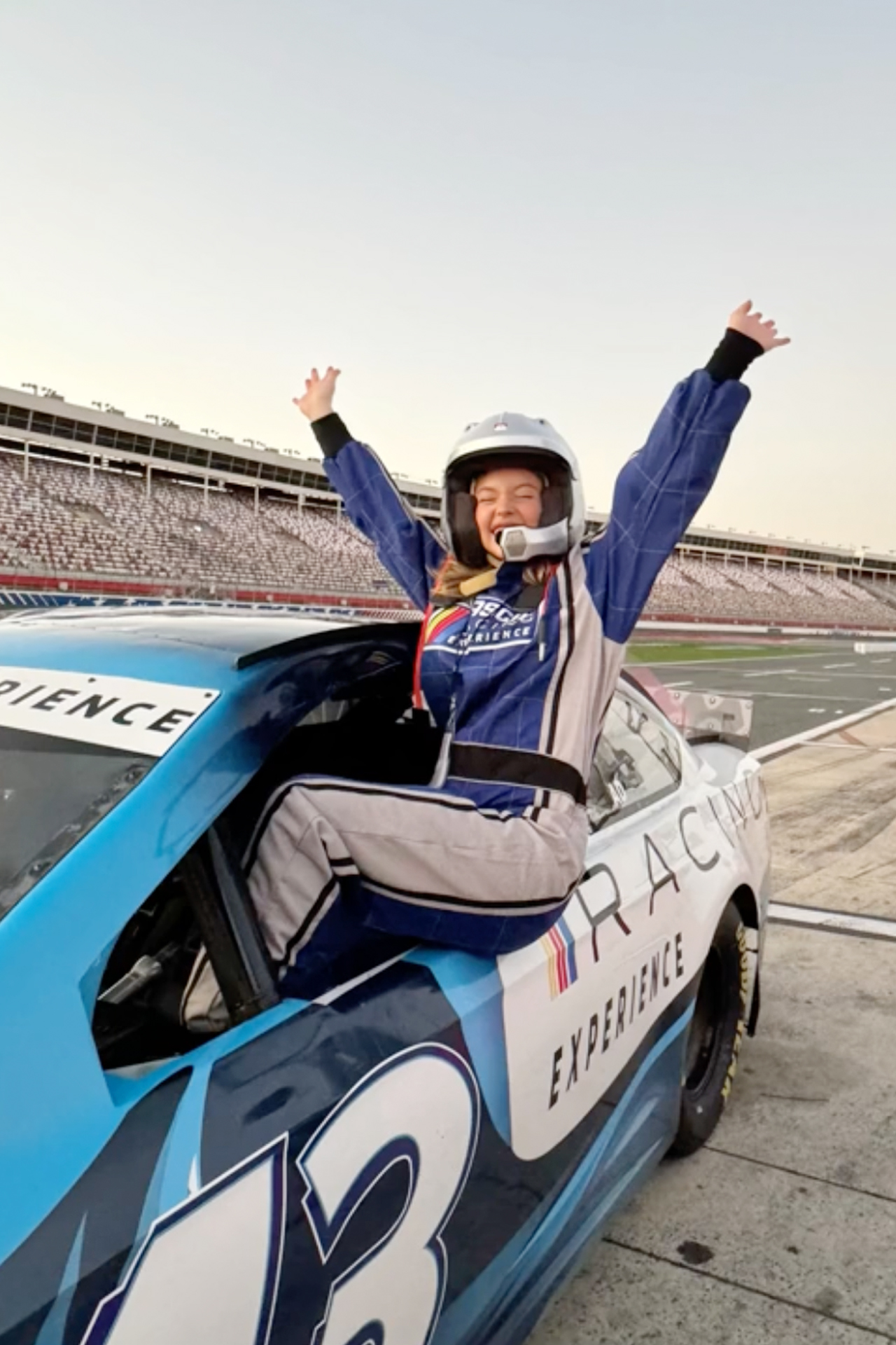 Sydney Sweeney shares her sheer delight as she leans out of a blue NASCAR car window with her arms triumphantly raised.