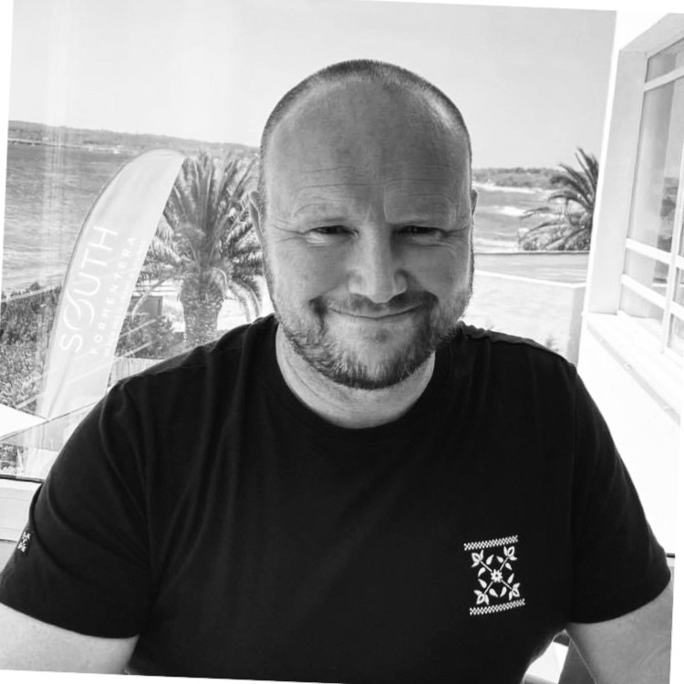 black and white headshot of male wearing black tshirt with sea view in background