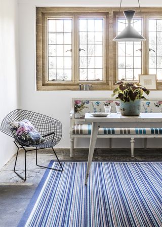 farmhouse cottage style dining area with blue stripe rug, floral vintage bench, white vintage table, modern black wire chair, retro pendant light, stone flooring