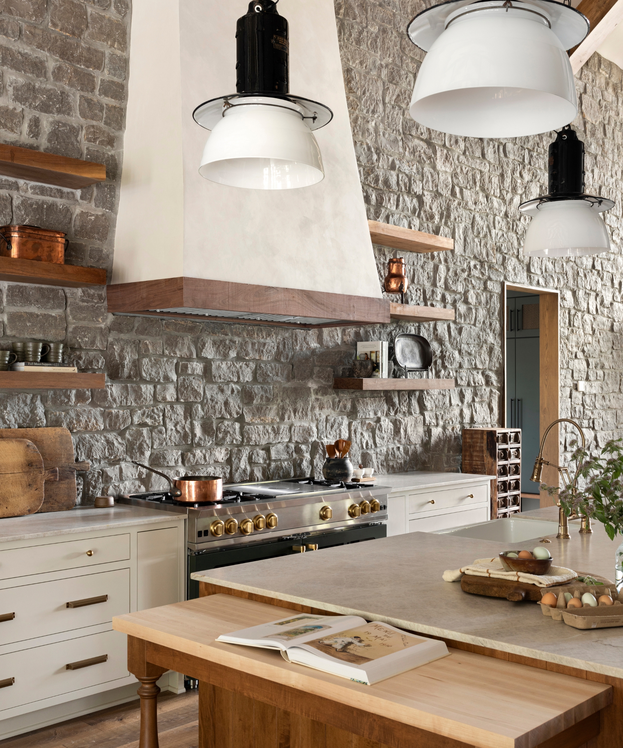 A rustic kitchen with exposed stone walls, white cabinets with oversized brass hardware, and wooden floating shelves