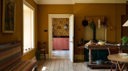 Medium kitchen with wooden floors, mismatched but stylish bench and wooden table. Through the open doorway, the yellow yellow wallpapered utility room with pink lower cabinets is visible in the background