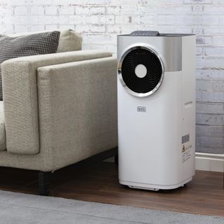 A white Black + Decker portable air conditioner sat on the livign room floor next to a beige sofa