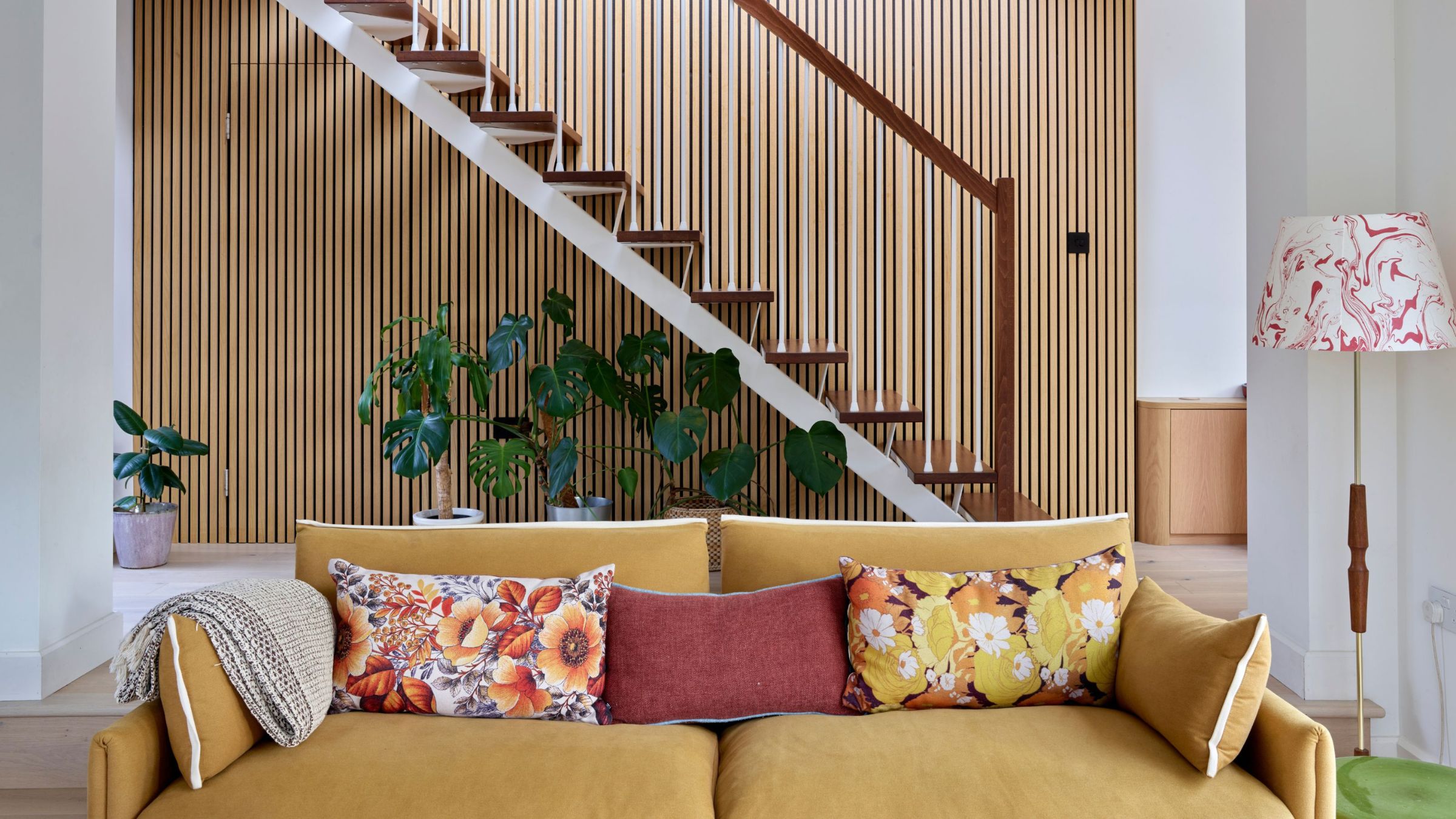 Yellow sofa with colourful floral cushions on it in front of a wood panelled staircase with plants underneath it 