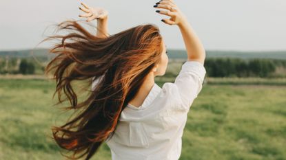 An image from the back of a woman in a green field tossing her hair back into the wind, wearing a white shirt 