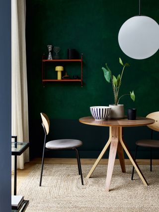 Dark green painted wall, compact round wood table, streamlined chair, and potted plant and mono stripe bowl on tabletop.