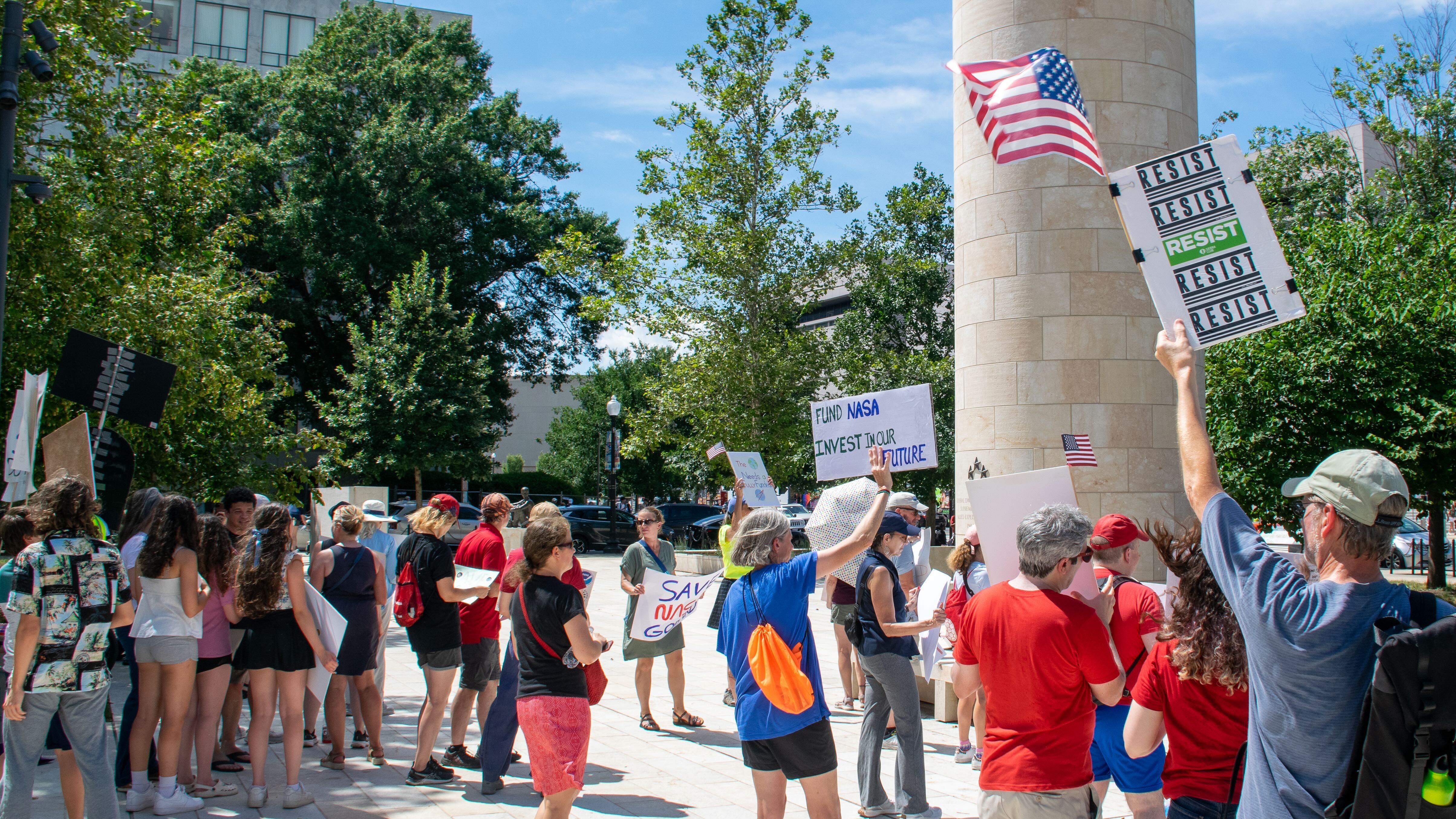 People hold signs outside on a sunny day in protest of NASA budget cuts, July 20, 2025.