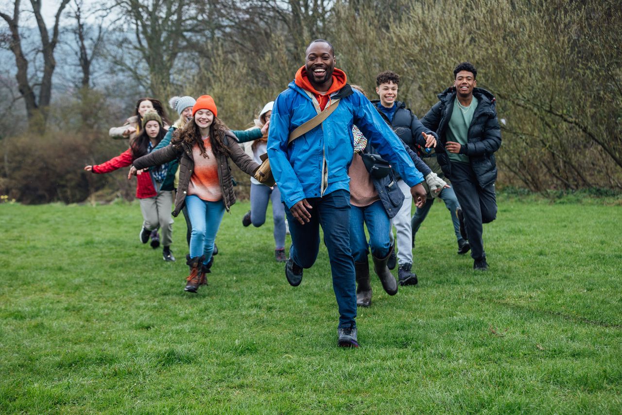 Our class having fun on a field trip. A wide shot of a large group of students and their two teachers wearing warm outdoor clothing on an overcast day. They are on a field trip and are laughing as they run towards the camera. 