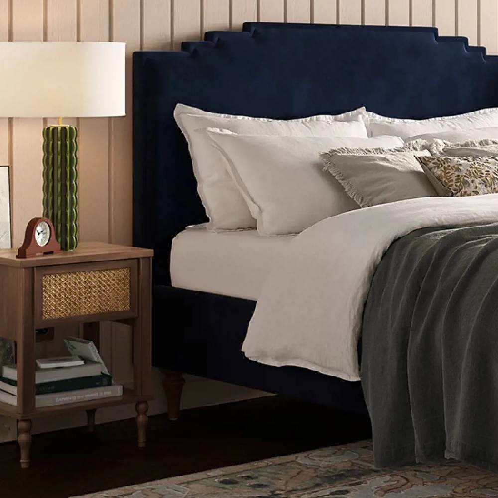 A modern, luxury bedroom with a navy blue headboard, cream panelling, and a wooden-rattan side table.