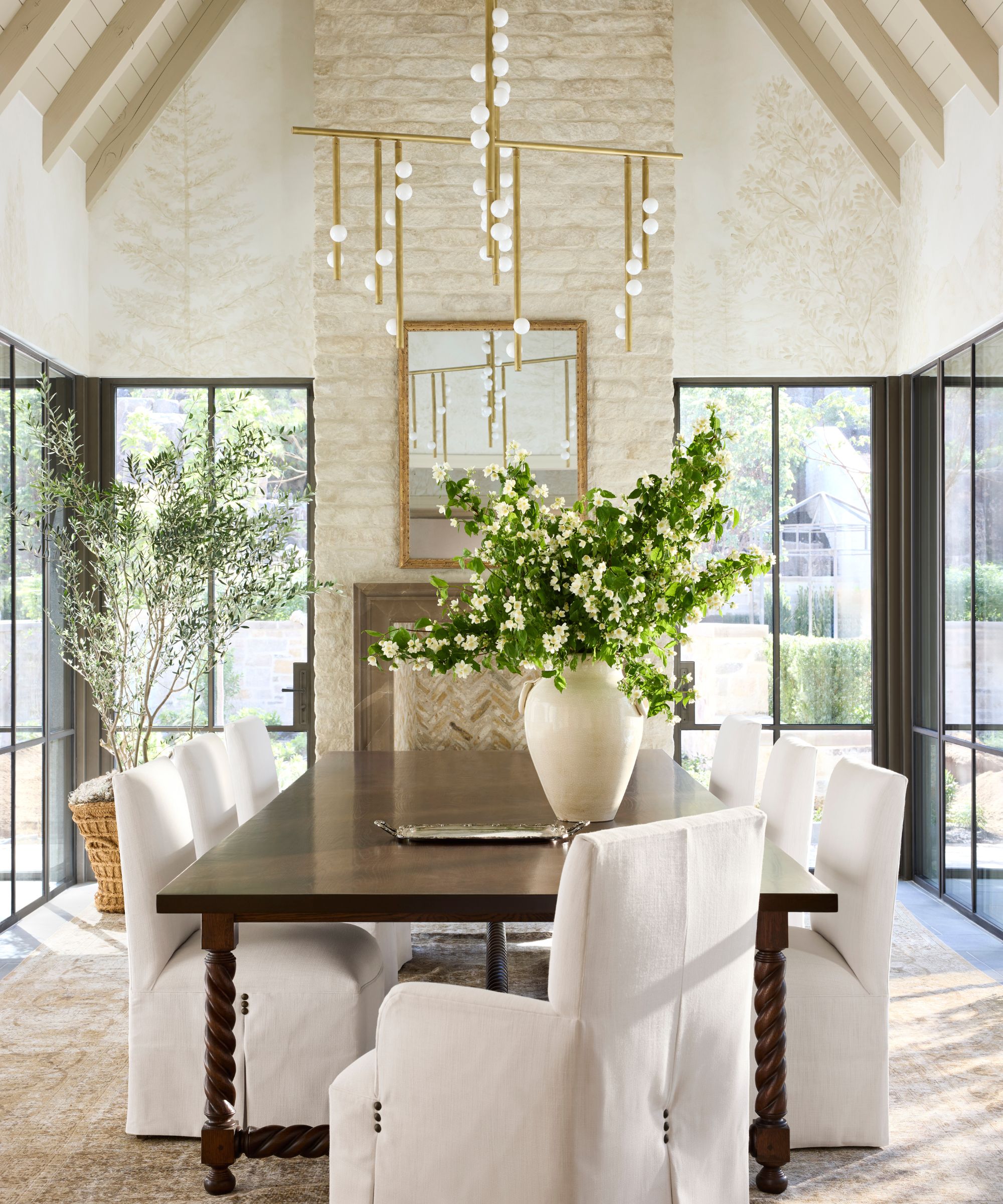 a large dining room with vaulted beamed ceiling, stone fireplace, glass walls and windows on either side and a large dark wooden table with twisted legs and linen chairs