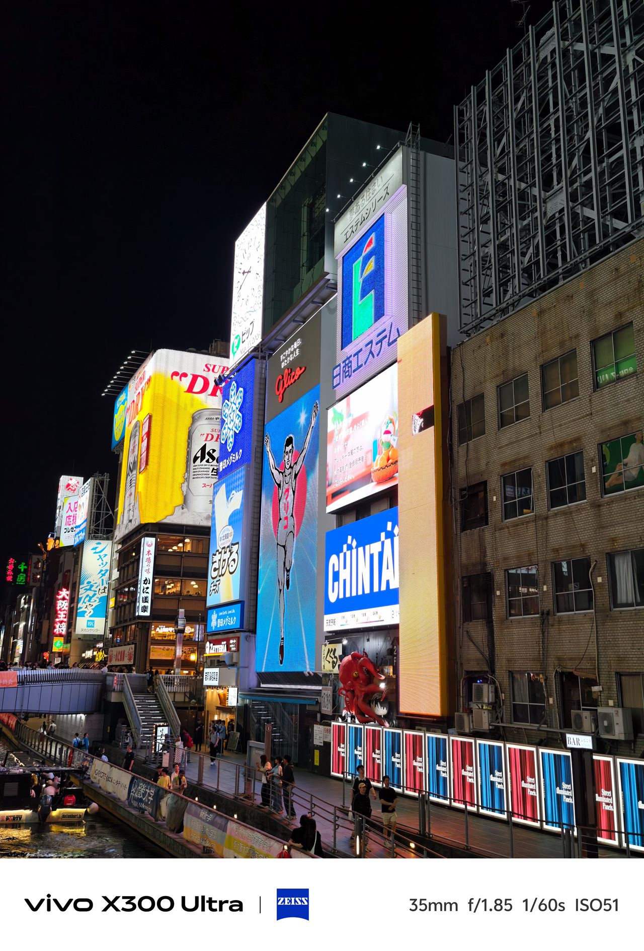 A close-up view of towering neon billboards, including the iconic running man sign, alongside a canal walkway at night.