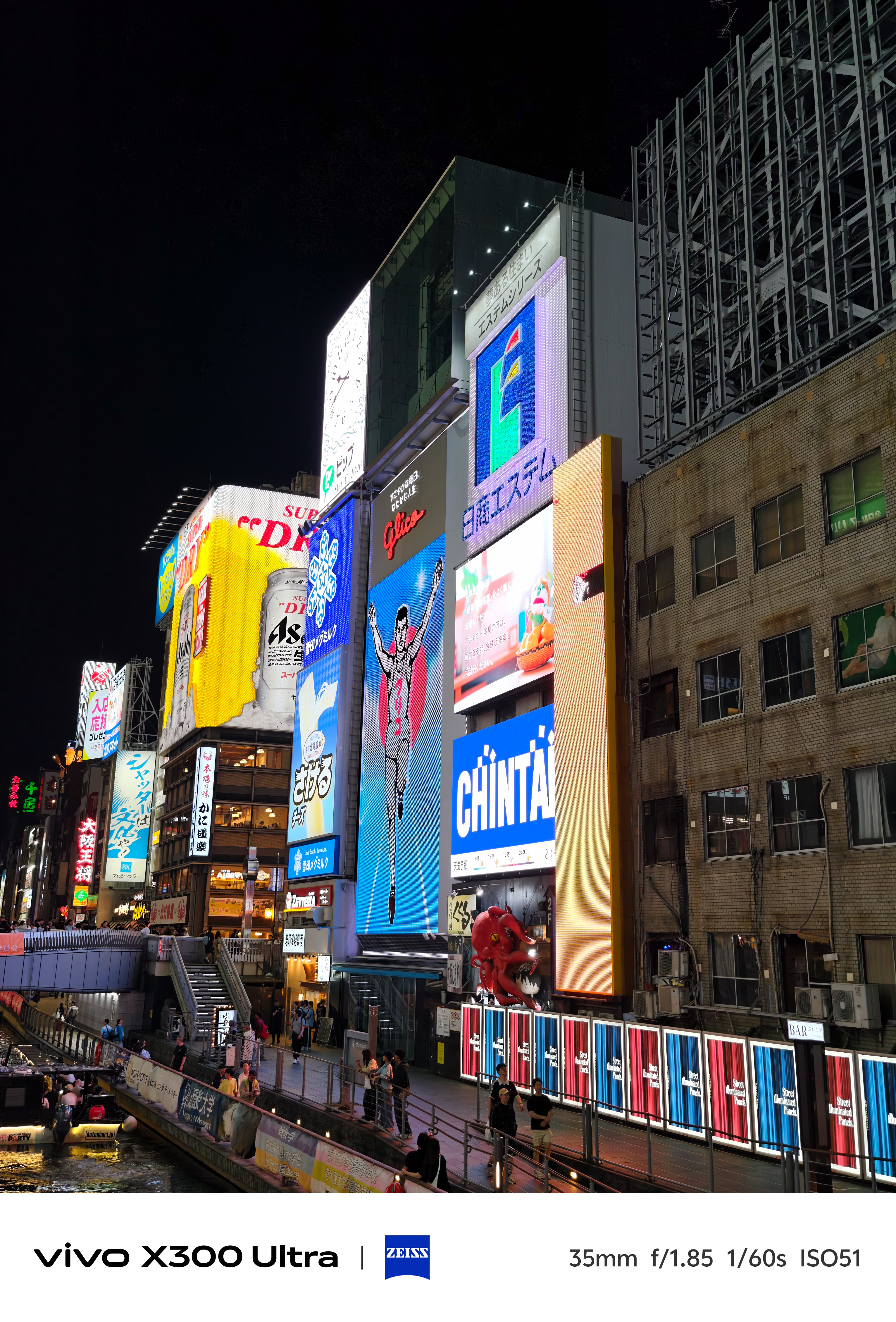 A close-up view of towering neon billboards, including the iconic running man sign, alongside a canal walkway at night.