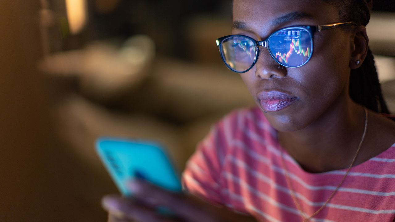 A stock graph is reflected in the glasses of a woman looking at her phone.