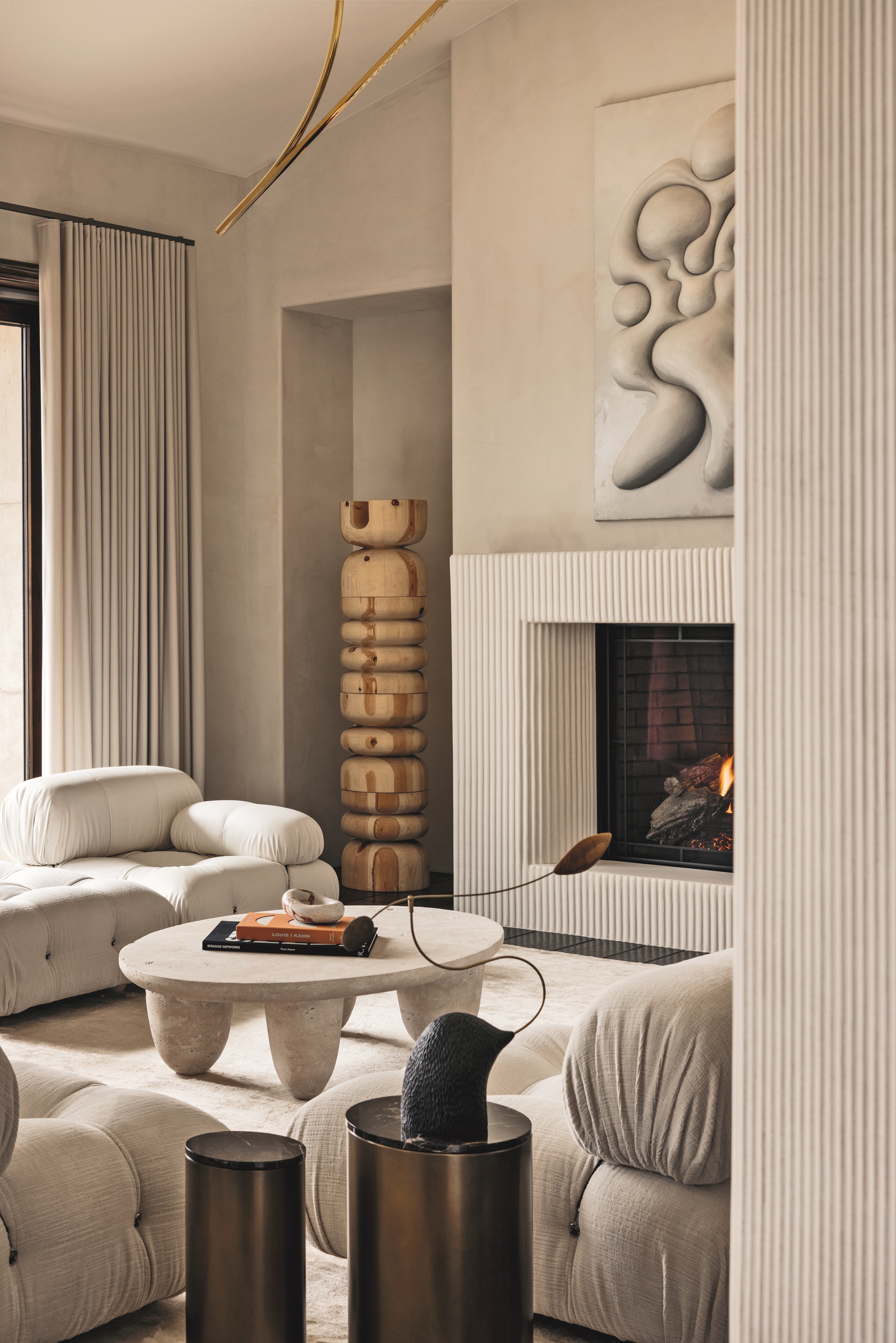 beige living room with fluted wall panels and fireplace surround, sculptural totem in the corner, beige blinds, stone coffee table, beige sofas, beige rugs, bronze side tables with sculptural decor