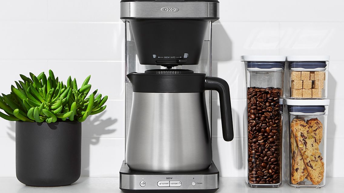 OXO Brew 8-Cup Coffee Maker on a kitchen countertop with a houseplant, and storage boxes of coffee beans and sugar