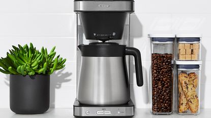 OXO 8-Cup Coffee Maker on a kitchen countertop with a houseplant, and storage boxes of coffee beans and sugar