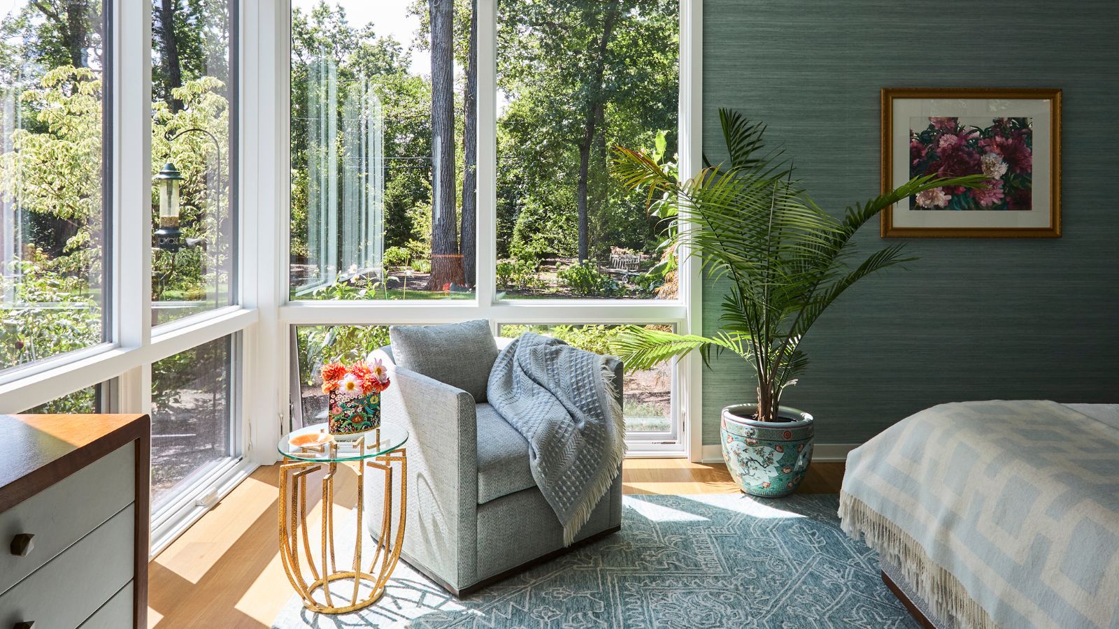 Bedroom with gray armchair with gray throw over it, large plant in vase, glass side table, large windows revealing outdoor area. Pictured to the right is the corner of a bed with a blue and white patterned rug.