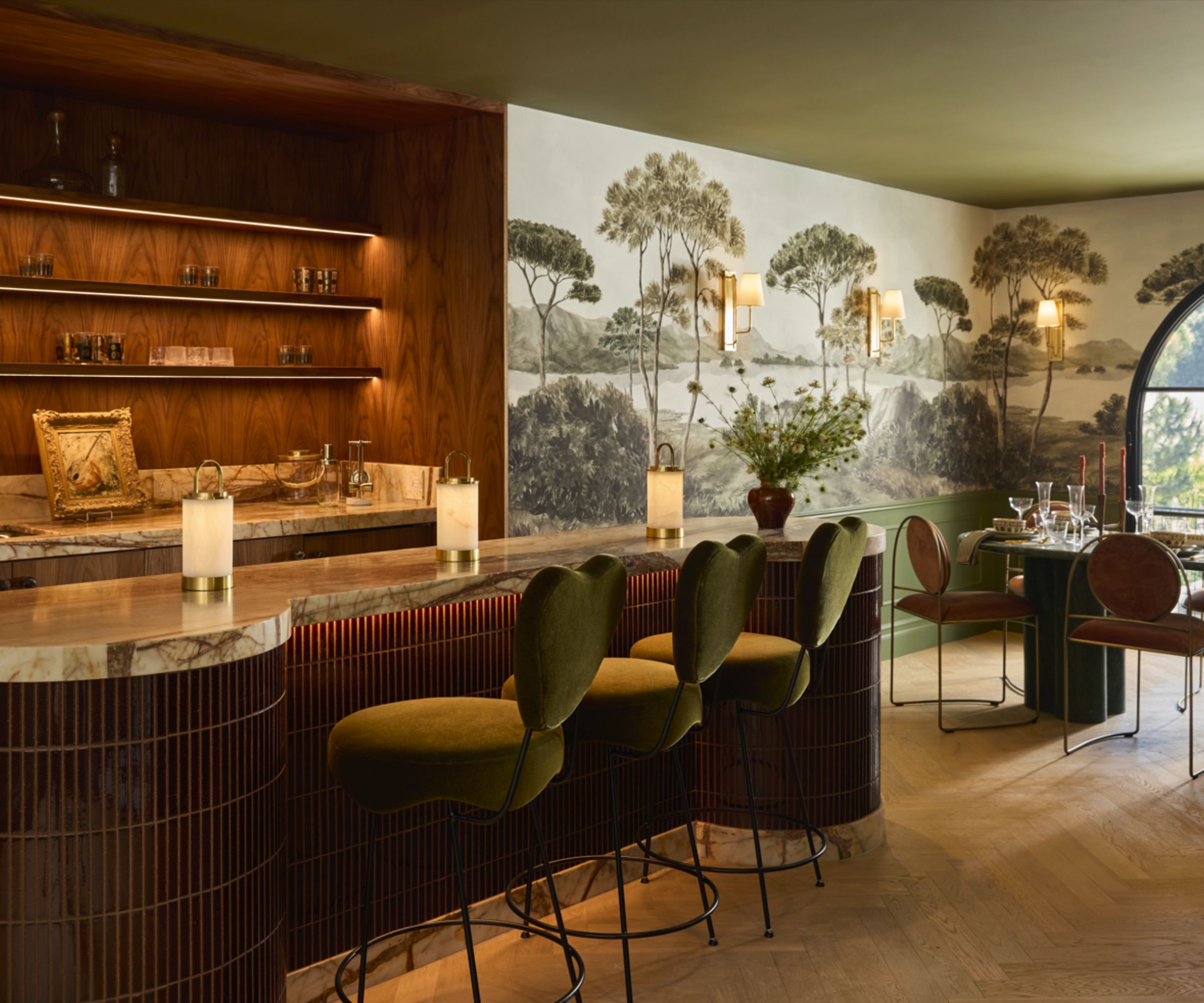 Luxurious home bar set-up with wooden shelving, marble countertop, cuved kitchen island with dark wood base, marble countertop, mural to the right-hand side in dining area with table and chairs