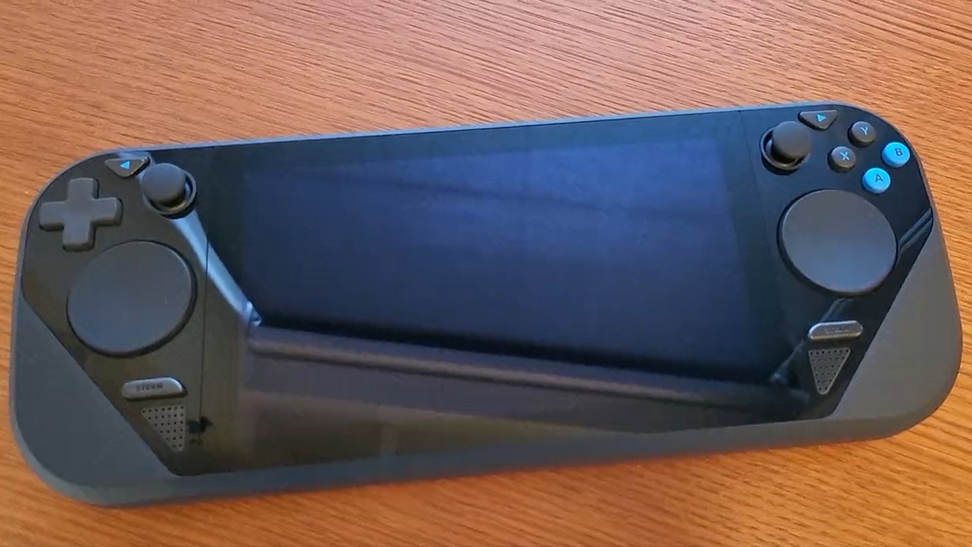Prototype Steam Deck with rounded trackpads, blue accents, smaller analogue sticks on top of a wooden table.