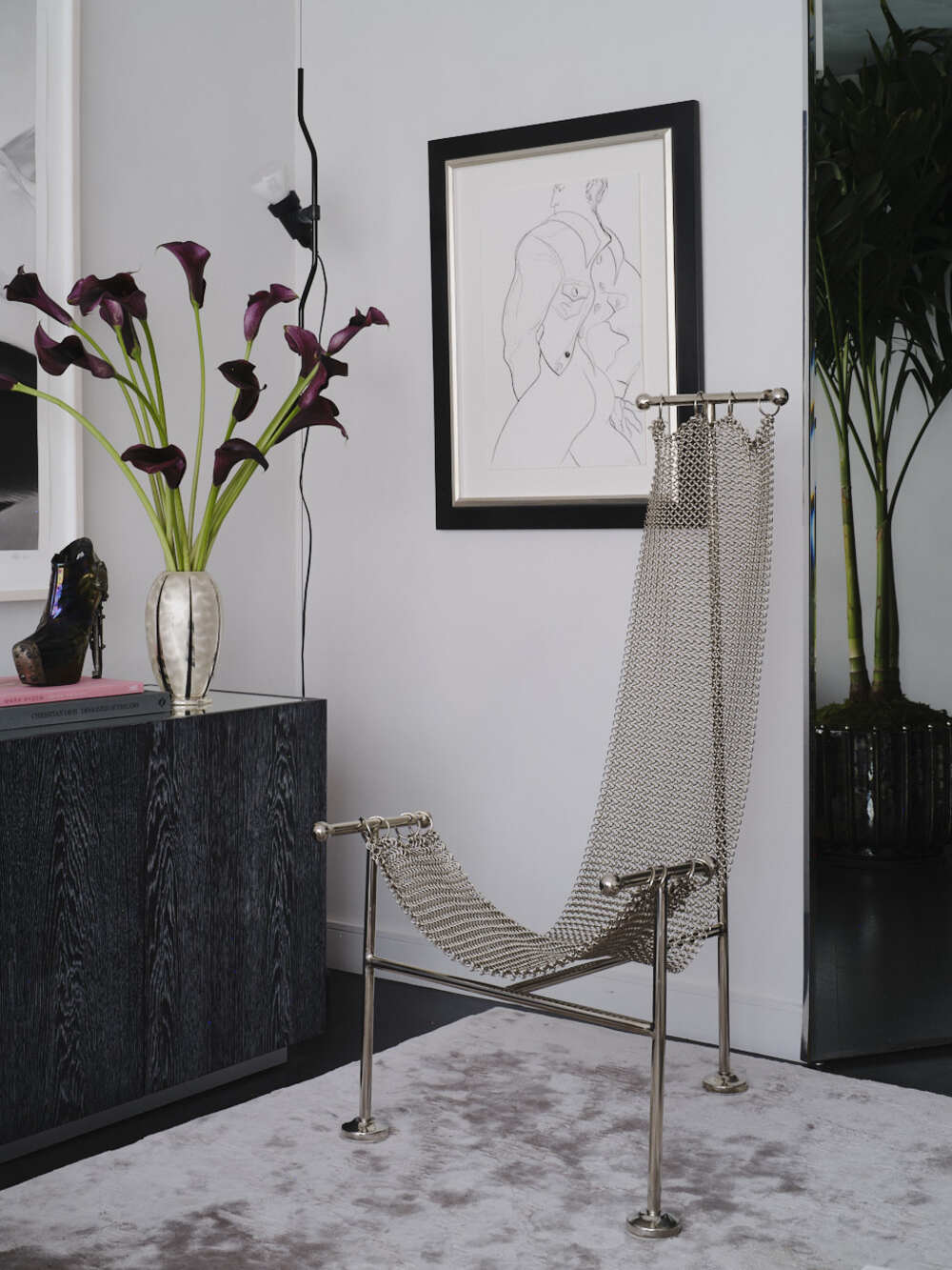 a chainmail chair int he corner of a modern living room with a black console styled with vase of flowers, an artwork on the wall, and a silk carpet