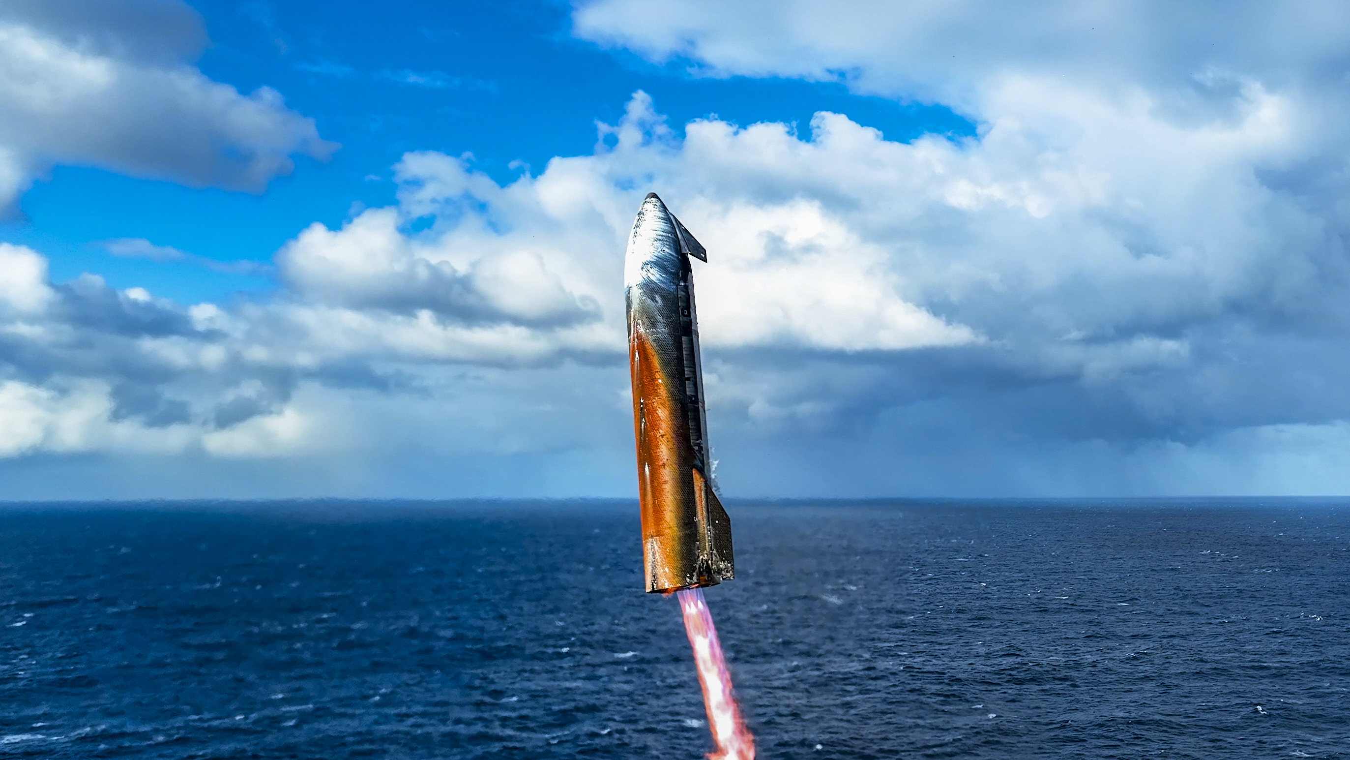 a burnt-looking silver rocket comes down for an ocean splashdown beneath cloudy blue skies